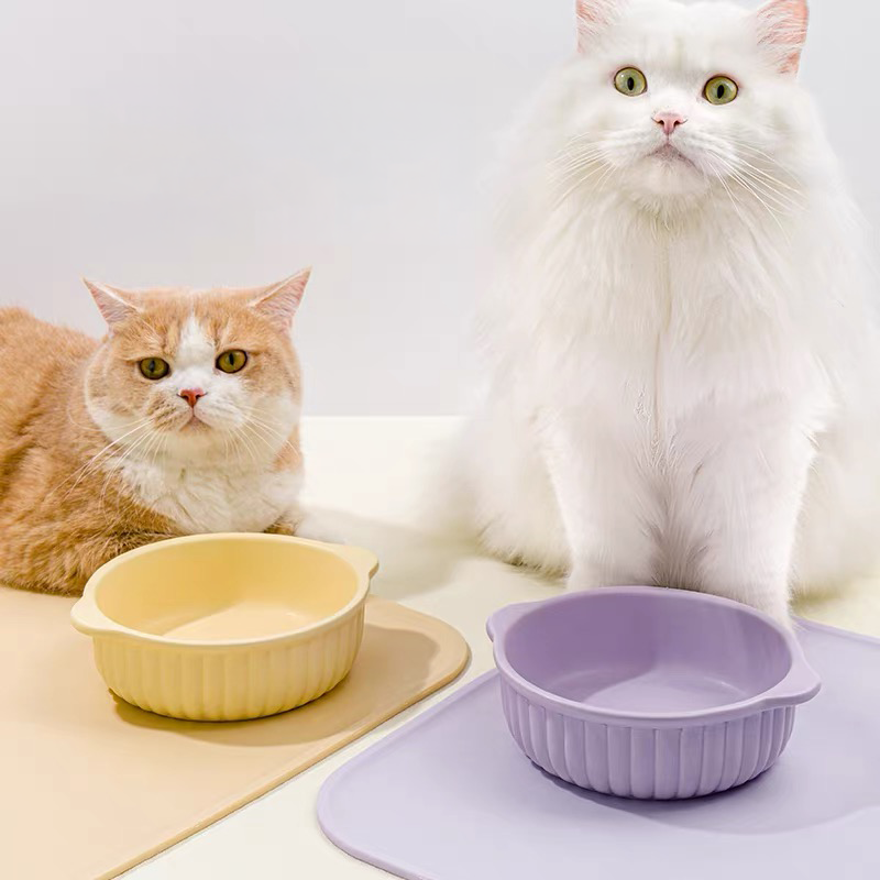 Two adorable fluffy cats sitting next to the yellow and purple Mango Dual-Ear ceramic bowls, which are placed on matching silicone feeding mats.