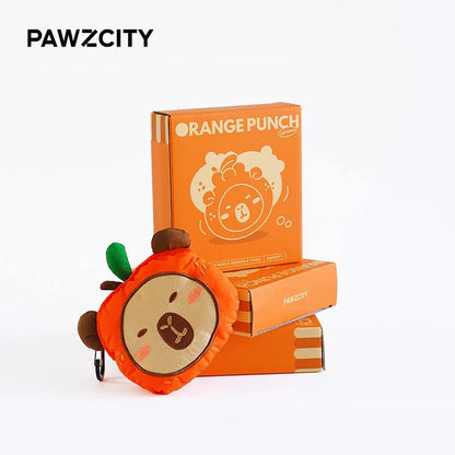 The PAWZCITY plush Capybara waste bag dispenser leaning against its matching vibrant orange packaging boxes labeled "ORANGE PUNCH CAPYBARA".