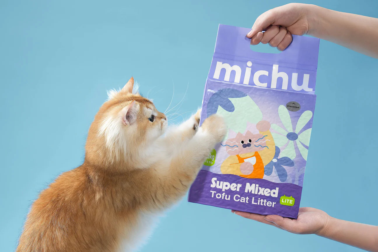 A playful golden cat reaching up with its paws toward a bag of Michu Mixed Tofu Cat Litter Lite held by a person, showing the pet&