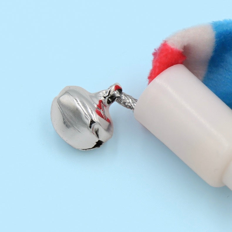 Close-up detail view of the metal bell attached to the base of a cat teaser wand, against a light blue background. The small, shiny silver metal bell hangs from a braided cord on a white plastic cap. Part of a multi-colored plush ribbon is visible, confirming safe claw and bite play materials that build trust and prevent injury.
