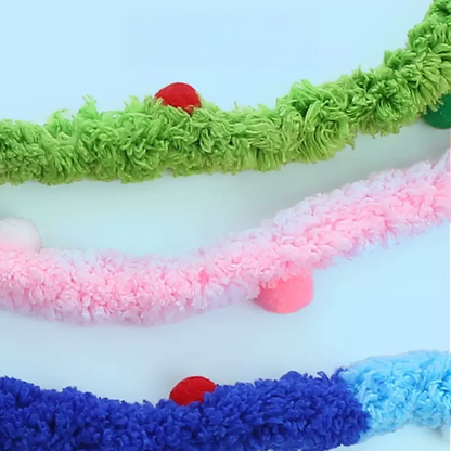 Close-up of three long, colorful plush cat teaser wand ribbons resting on a light blue background. The top ribbon is green with red and green pom-poms, the middle ribbon is pink with pink and white pom-poms, and the bottom ribbon is blue and light blue with a red pom-pom. These long, wavy tails mimic slithering prey for hunting instincts.