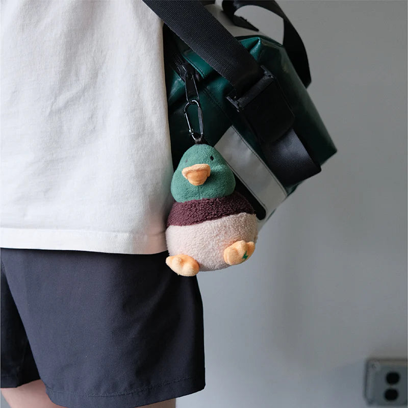 A person carrying a green and black travel bag with the PAWZCITY plush duck waste bag dispenser attached to the strap as a cute and functional charm.