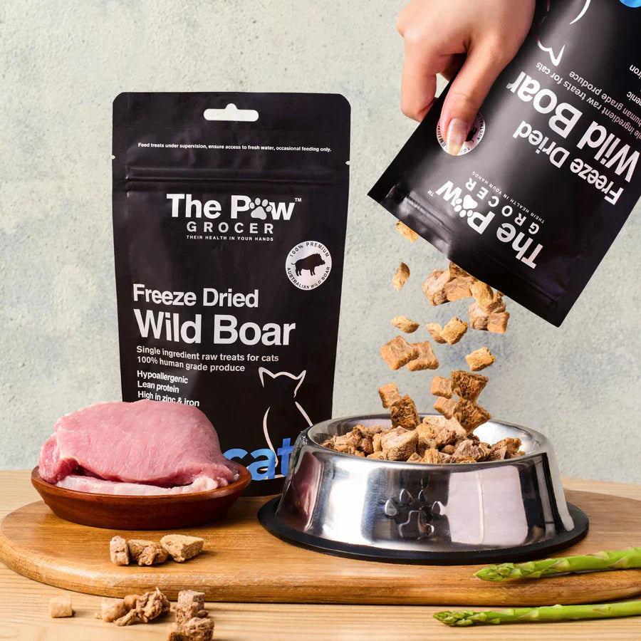 A hand pouring 100% Australian freeze-dried wild boar cat treats from the pouch into a silver pet bowl, showcasing the natural, raw cubed texture.