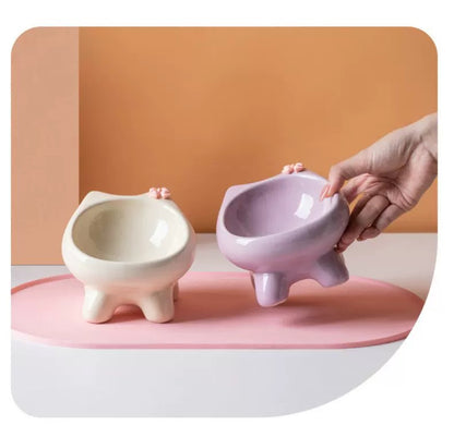 A hand gently lifting a purple cat-ear ceramic bowl next to a white one on a pink mat, illustrating the stable, elevated paw-shaped base.