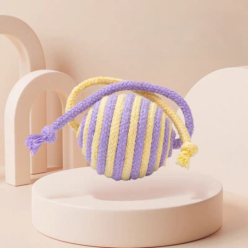 A close-up view of a purple and yellow pastel striped sisal cat toy ball resting on a minimalist beige podium.