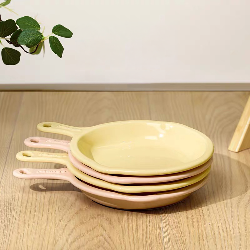 A neat stack of pastel yellow and pink skillet-shaped ceramic pet plates resting on a wooden floor, showcasing their space-saving and home-cooked aesthetic design.