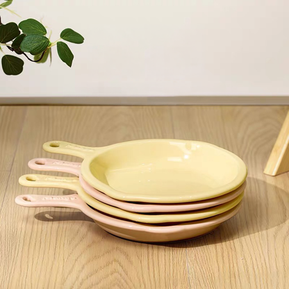 A neat stack of pastel yellow and pink skillet-shaped ceramic pet plates resting on a wooden floor, showcasing their space-saving and home-cooked aesthetic design.