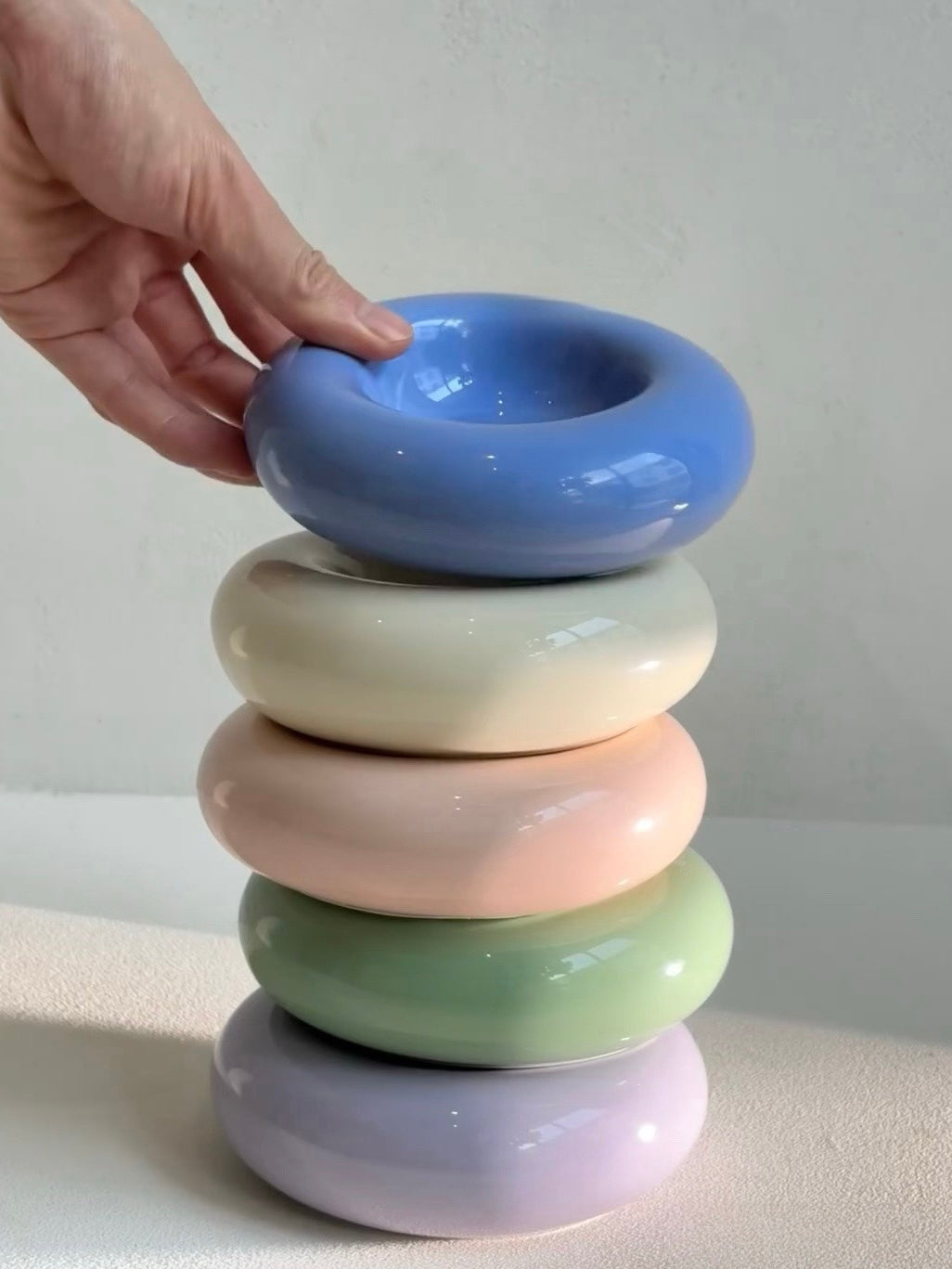 A hand stacking five ceramic bagel pet bowls in pastel colors: blue, cream, peach, mint green, and lilac, highlighting their stackable design and aesthetic appeal.