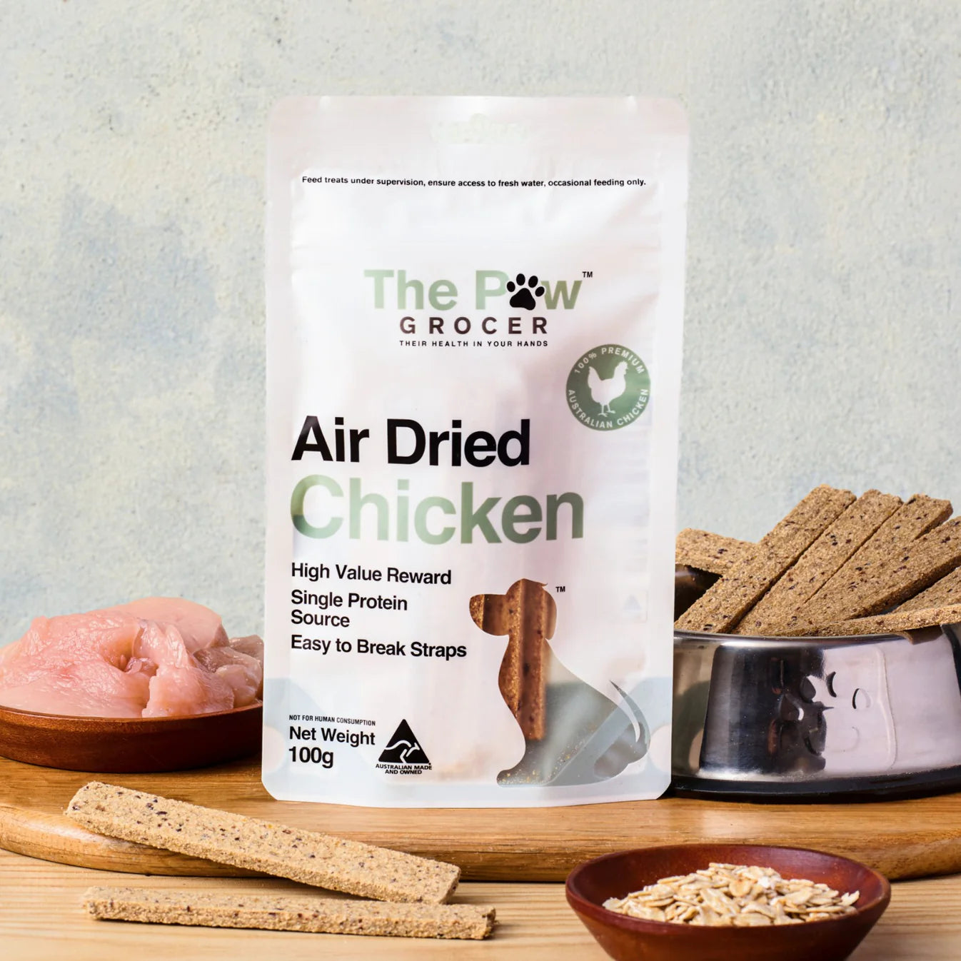 Detailed showcase of The Paw Grocer Air Dried Chicken packaging on a wooden board next to fresh chicken, oats, and a bowl of dried straps.