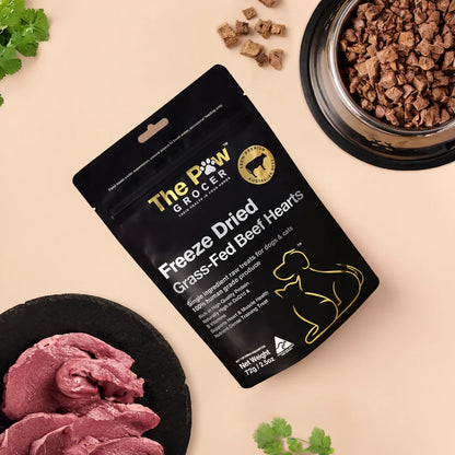 A top-down view of The Paw Grocer Freeze-Dried Grass-Fed Beef Hearts pouch surrounded by fresh raw beef heart and a silver bowl filled with small, park-ready cubed training treats.