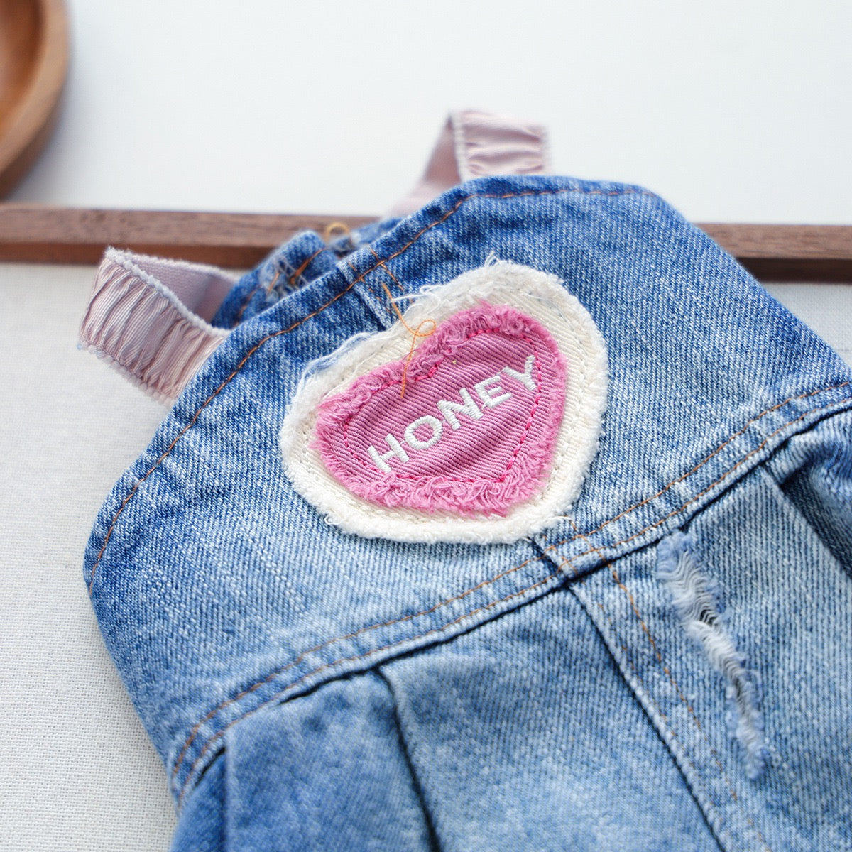 Macro close-up of the Honey Heart denim dog dress, detailing the pink embroidered heart patch, neat stitching, and subtle distressed vintage wash edges.
