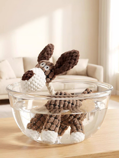 The brown dog Bubble Farm plush toy completely submerged in a clear glass bowl of water, demonstrating that it is 100% fully washable and colorfast.