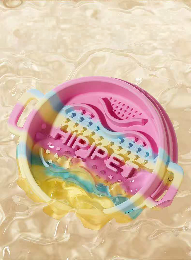 A pink and yellow pastel swirl silicone slow feeder bowl being splashed with clear water, demonstrating its waterproof, easily washable, and food-grade material.