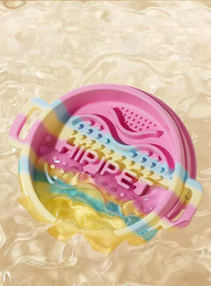 A pink and yellow pastel swirl silicone slow feeder bowl being splashed with clear water, demonstrating its waterproof, easily washable, and food-grade material.
